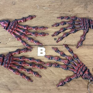 May include: Four red and black skeleton hand keyrings with silver key rings. The keychains are arranged in a circle on a wooden surface. The letter "B" is visible in the centre of the image.