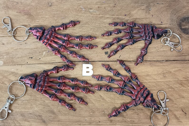 May include: Four red and black skeleton hand keyrings with silver key rings. The keychains are arranged in a circle on a wooden surface. The letter "B" is visible in the centre of the image.
