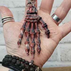 May include: A red and black skeleton hand keyring. The hand is made of plastic and has a detailed bone structure. The keyring is attached to a silver ring.