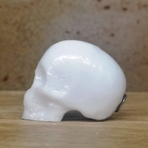 May include: A white ceramic skull figurine with a smooth, glossy finish. The skull is facing to the side and has a small metal ring on the back.