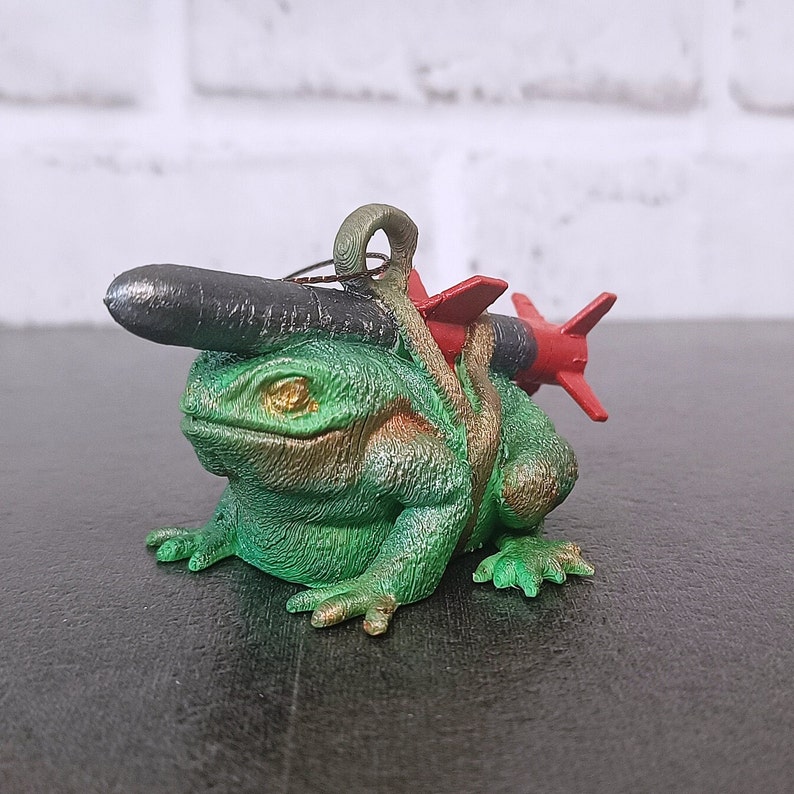 May include: A green frog figurine with a black and red rocket strapped to its back. The frog has a textured, bumpy surface and a wide, toothy grin.