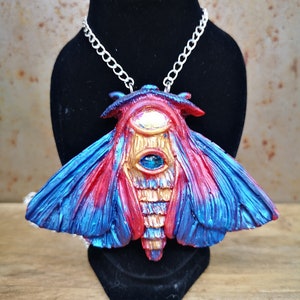 May include: A silver chain necklace with a large, red, blue, and gold moth pendant. The moth has a large, blue eye in the center of its body.