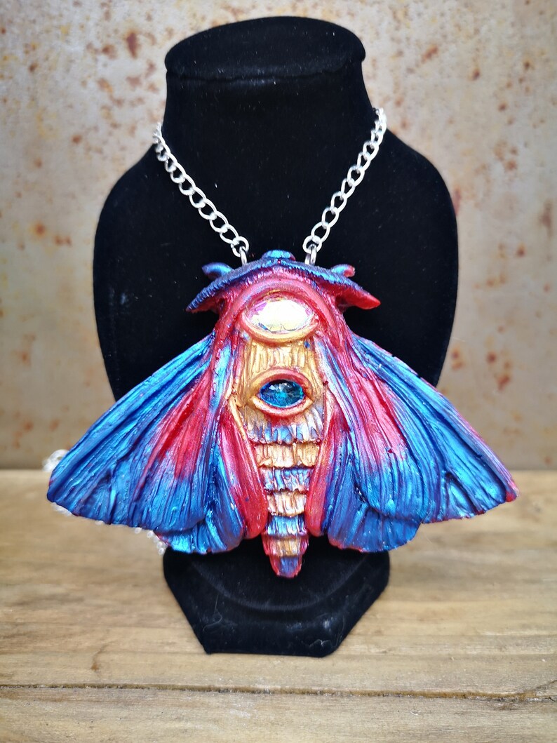 May include: A silver chain necklace with a large, red, blue, and gold moth pendant. The moth has a large, blue eye in the center of its body.