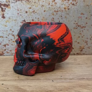 May include: A red and black skull-shaped planter with a marbled design. The planter is made of ceramic and has a smooth, glossy finish.