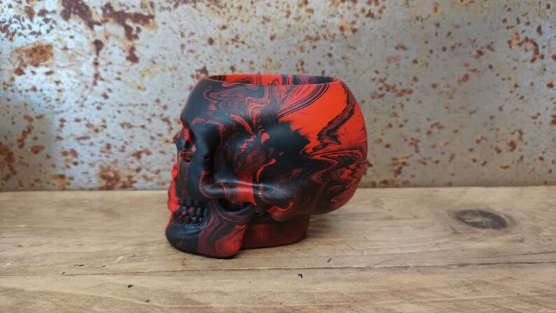 May include: A red and black skull-shaped planter with a marbled design. The planter is made of ceramic and has a smooth, glossy finish.