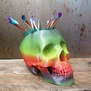 May include: A rainbow-colored skull-shaped ceramic holder with a variety of colorful paintbrushes sticking out of the top.