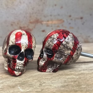 May include: Two white ceramic skull drawer pulls with red stripes. The skulls have black eye sockets and are detailed with a textured surface.
