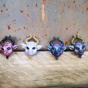 Puede incluir: Cuatro peque&ntilde;as figuritas de resina de color de criaturas con cuernos pintadas de negro, dorado, plateado y morado. Las figuritas est&aacute;n dispuestas en fila sobre una superficie de madera.