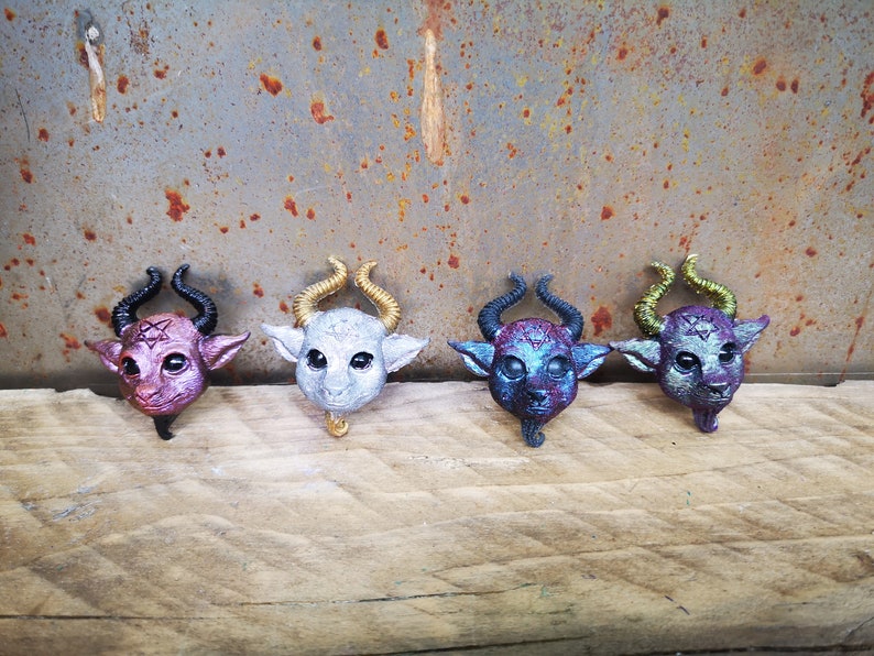 Puede incluir: Cuatro peque&ntilde;as figuritas de resina de color de criaturas con cuernos pintadas de negro, dorado, plateado y morado. Las figuritas est&aacute;n dispuestas en fila sobre una superficie de madera.