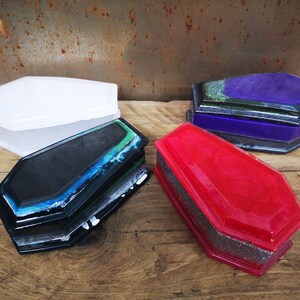 May include: Four small coffin-shaped boxes made of resin. The boxes are in various colors, including white, black, purple, and red. Each box has a glittery finish.