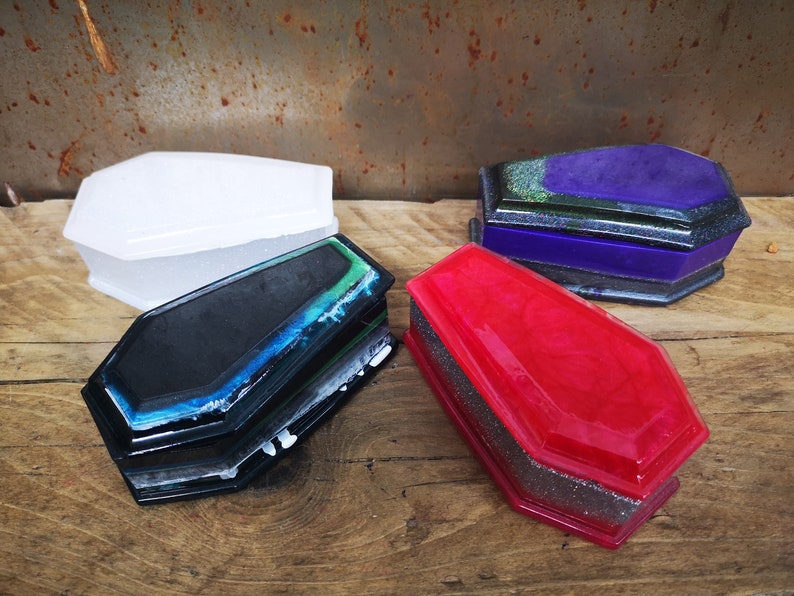 May include: Four small coffin-shaped boxes made of resin. The boxes are in various colors, including white, black, purple, and red. Each box has a glittery finish.