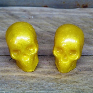 May include: Two gold-colored skull-shaped figurines with a shiny, metallic finish. The skulls are facing forward and have a detailed design.