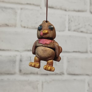 Puede incluir: Un adorno de pájaro marrón y dorado con un babero rojo y rosa. El pájaro tiene ojos grandes y redondos y un pico pequeño. Está colgado de una cuerda marrón.