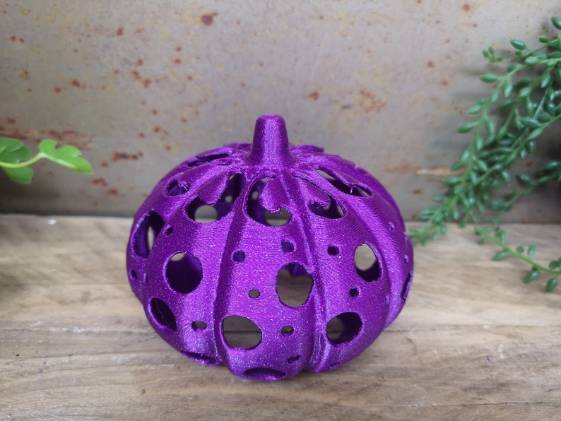 May include: A purple, glittery, hollow pumpkin with a decorative design. The pumpkin is made of plastic and has a round shape.