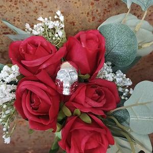May include: A bouquet of red roses with a silver skull charm in the centre. The roses are surrounded by small white flowers and green leaves.