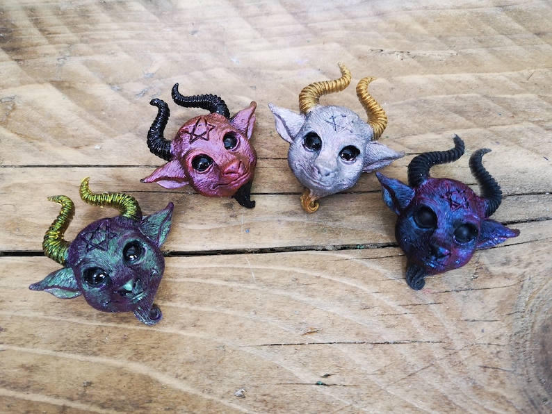 Puede incluir: Cuatro peque&ntilde;as figuritas de resina de color de criaturas con cuernos con s&iacute;mbolos de pentagrama en la frente. Las figuritas est&aacute;n pintadas en tonos de morado, azul, verde y plateado.