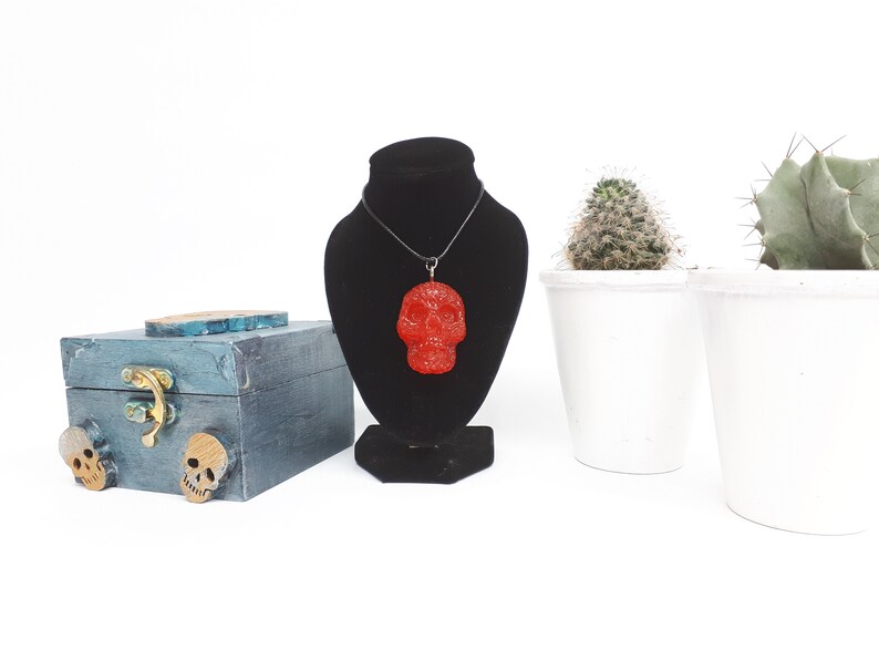 May include: A red glitter skull pendant necklace on a black cord, displayed on a black velvet stand. The necklace is next to a small wooden box with two skull accents and two potted cacti.