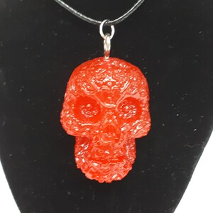 May include: A red resin skull pendant on a black cord necklace. The skull has a detailed, textured surface.