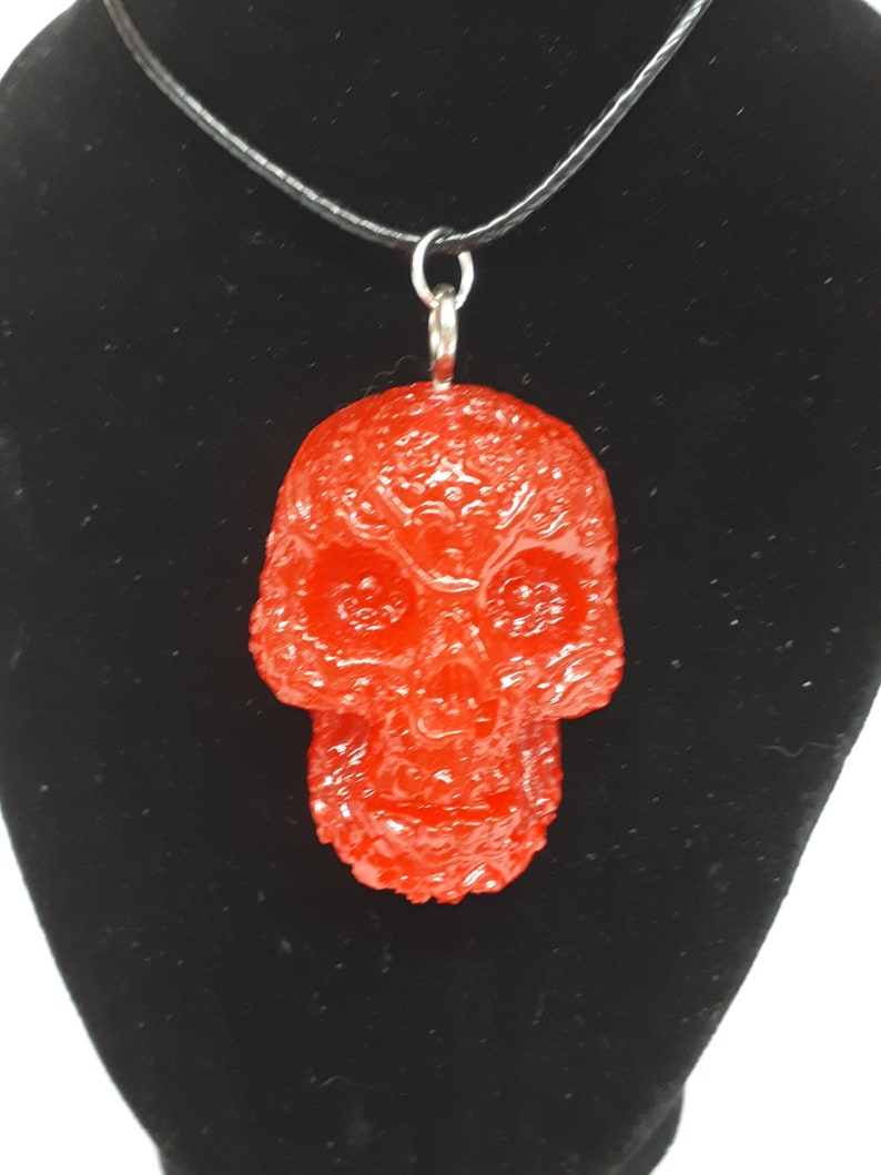 May include: A red resin skull pendant on a black cord necklace. The skull has a detailed, textured surface.