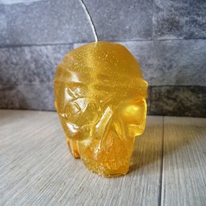 May include: A gold skull-shaped candle with a glitter finish. The candle is on a wooden surface.