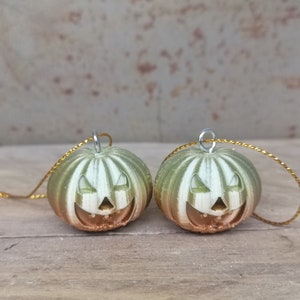 K&ouml;nnte beinhalten: Zwei gr&uuml;ne und goldene K&uuml;rbis-Anh&auml;nger mit einem Jack-o'-Lantern-Gesicht. Jeder Anh&auml;nger hat eine goldene Schnur.
