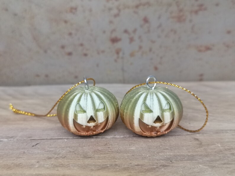 K&ouml;nnte beinhalten: Zwei gr&uuml;ne und goldene K&uuml;rbis-Anh&auml;nger mit einem Jack-o'-Lantern-Gesicht. Jeder Anh&auml;nger hat eine goldene Schnur.