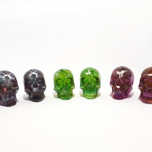 May include: Six resin skull ornaments in a row, each with a different colour. The colours include green, purple, and black. The skulls have a glossy finish and are set against a white background. A unique decorative item.