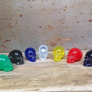 May include: Eight small glass skull figurines in various colors, including green, black, blue, clear, yellow, and red. The skulls are arranged in a row on a wooden surface.