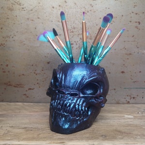 May include: A black and teal skull-shaped ceramic makeup brush holder with a variety of teal and purple makeup brushes sticking out of the top.