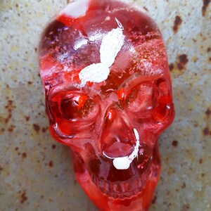 May include: A red glass skull with white paint details, resembling a butterfly and a mustache. The skull is translucent and has a glossy finish.
