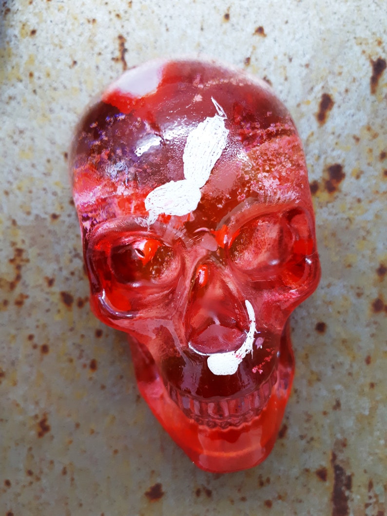 May include: A red glass skull with white paint details, resembling a butterfly and a mustache. The skull is translucent and has a glossy finish.