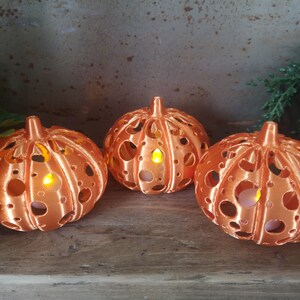 May include: Three orange pumpkin-shaped candle holders with a decorative cut-out design. Each holder has a small LED candle inside, casting a warm glow.