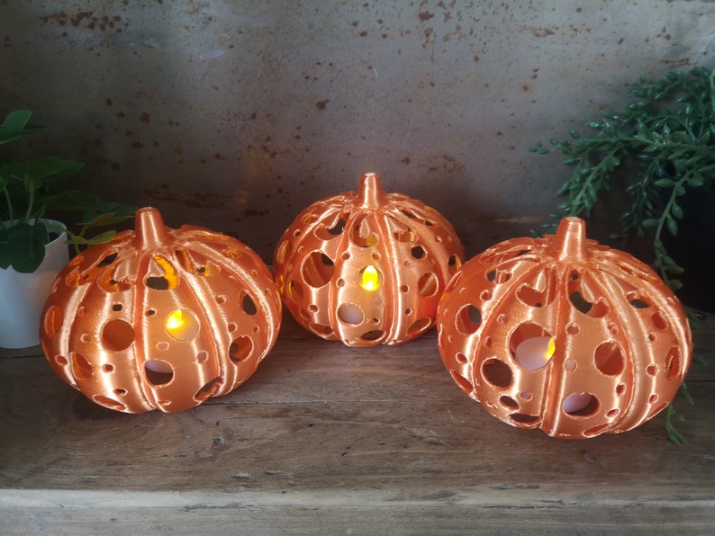 May include: Three orange pumpkin-shaped candle holders with a decorative cut-out design. Each holder has a small LED candle inside, casting a warm glow.