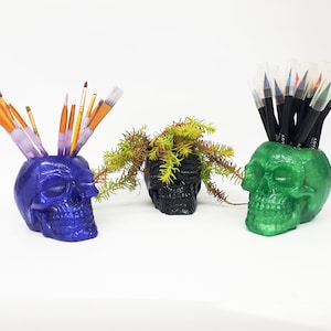 May include: Three skull-shaped planters, one purple, one black, and one green, with different items inside. The purple planter holds paintbrushes, the black planter holds a small plant, and the green planter holds colored markers.