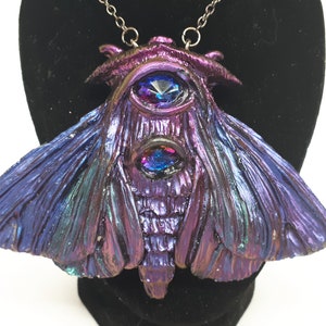 May include: A purple and blue iridescent moth pendant with two large, faceted, blue and purple gemstones for eyes. The pendant is on a silver chain.