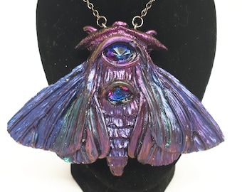 May include: A purple and blue iridescent moth pendant with two large, faceted, blue and purple gemstones for eyes. The pendant is on a silver chain.