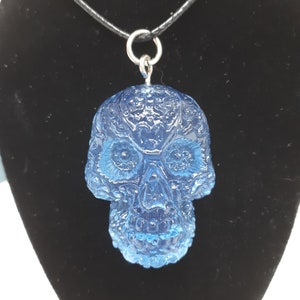 May include: A blue glass skull pendant necklace with intricate detailing. The pendant is attached to a black cord necklace.