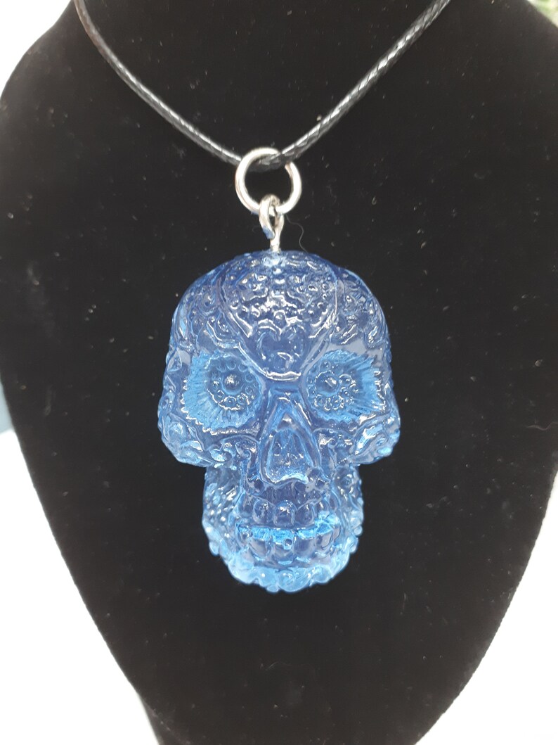 May include: A blue glass skull pendant necklace with intricate detailing. The pendant is attached to a black cord necklace.