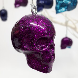 Puede incluir: Un adorno de calavera de purpurina morada que cuelga de una cuerda plateada. La calavera es detallada y tiene un acabado brillante.