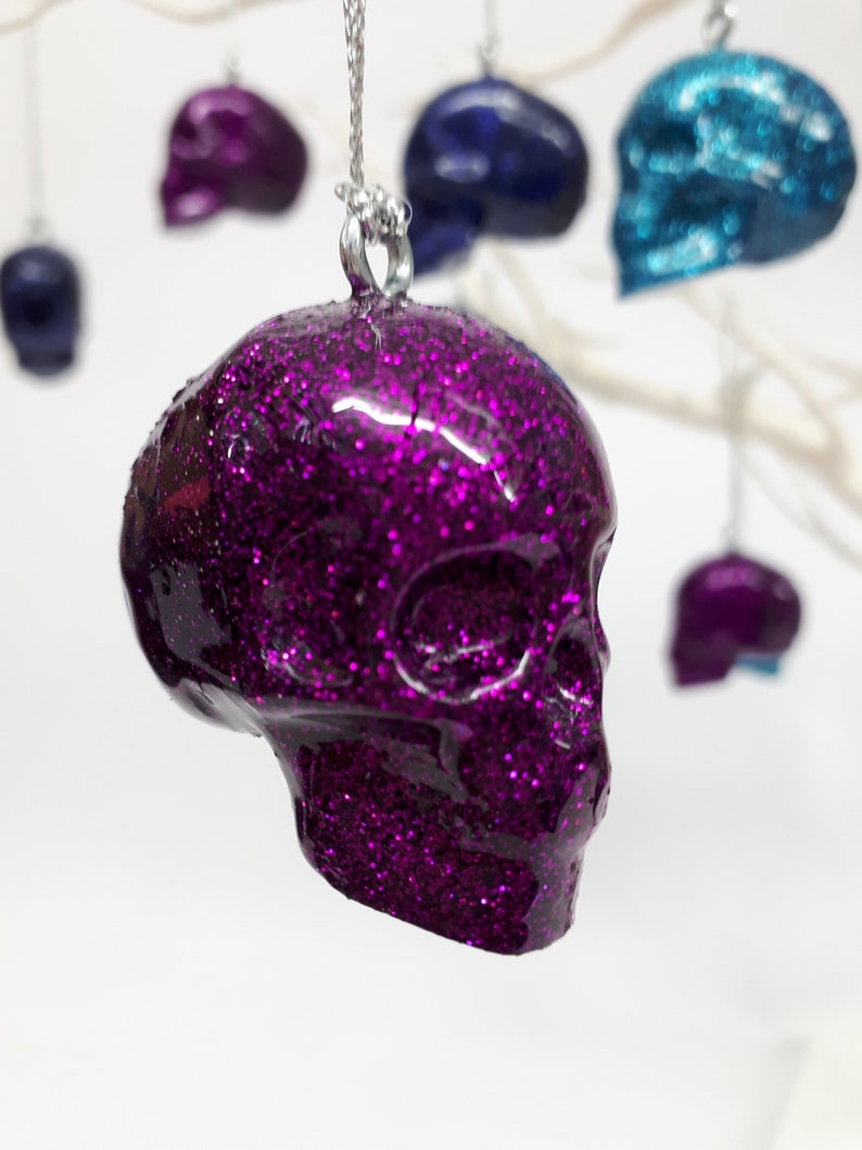 Puede incluir: Un adorno de calavera de purpurina morada que cuelga de una cuerda plateada. La calavera es detallada y tiene un acabado brillante.