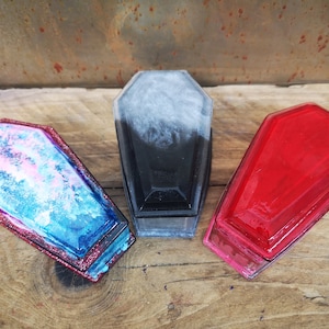May include: Three small coffin-shaped boxes made of resin. The boxes are decorated with different colors and patterns. One box is black with white swirls, one is red, and one is blue with pink and white swirls.