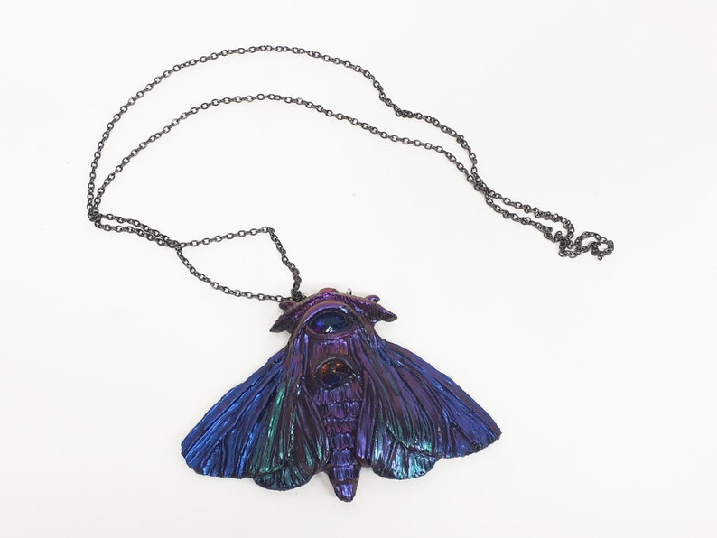 May include: A iridescent blue and purple moth pendant necklace with a black chain. The moth has a large eye on its head and intricate details on its wings.