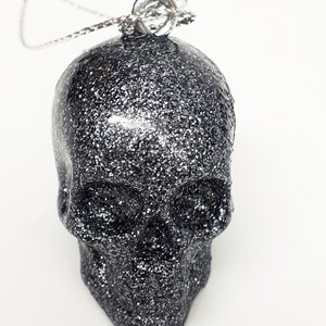 Puede incluir: Un colgante de calavera de purpurina plateada en una cadena plateada.