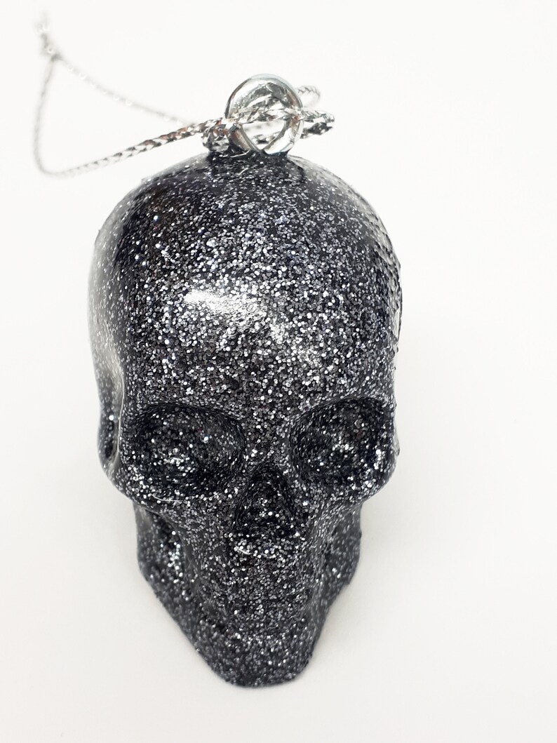 Puede incluir: Un colgante de calavera de purpurina plateada en una cadena plateada.