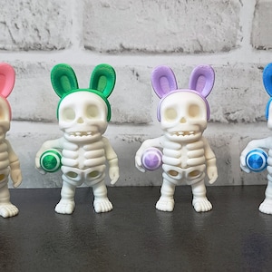 May include: Four white skeleton figurines with bunny ears and holding colourful Easter eggs. The skeletons are wearing pink, green, purple, and blue bunny ears. The eggs are pink, green, purple, and blue.