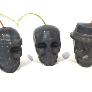 May include: Three black skull ornaments with gold and copper wire loops. The skulls have different facial features and one has a small black hat.