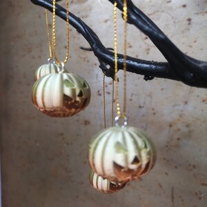 K&ouml;nnte beinhalten: Vier gr&uuml;ne und braune K&uuml;rbis-Ornamente mit Jack-o'-Lantern-Gesichtern, die an goldenen Ketten an einem schwarzen Ast h&auml;ngen.