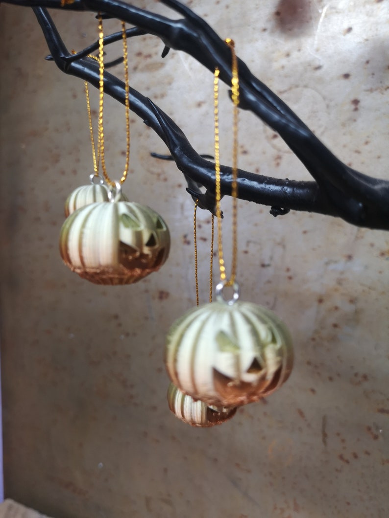 K&ouml;nnte beinhalten: Vier gr&uuml;ne und braune K&uuml;rbis-Ornamente mit Jack-o'-Lantern-Gesichtern, die an goldenen Ketten an einem schwarzen Ast h&auml;ngen.