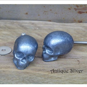 May include: Two antique silver skull drawer pulls with a screw and nut. The skull pulls are on a wooden surface.