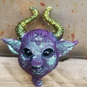 Puede incluir: Una figurita de resina iridiscente de color morado y verde de un peque&ntilde;o demonio con cuernos dorados y un pentagrama en la frente.